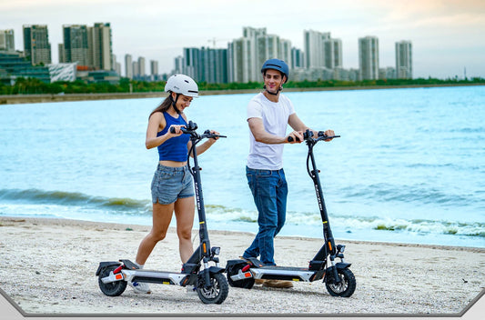 A man and a woman are walking along a beach, pushing a Kukirin electric scooter.
